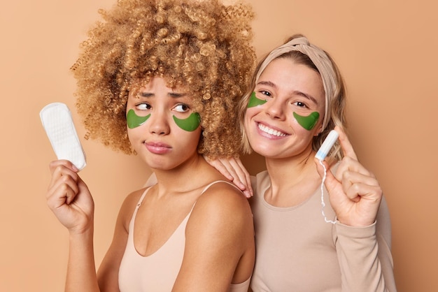 Photo of two women friends pose with hygiene products choose between sanitary napkin and tampon apply hydrogel patches under eyes undergo beauty procedures stand closely to each other indoors