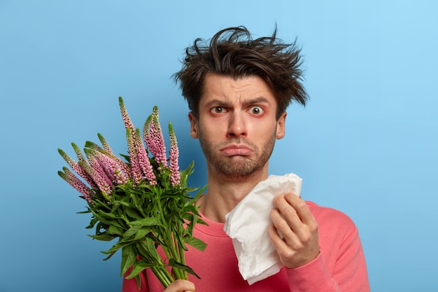 Photo of unhappy sick young man has problems with health, rubs nose with paper tissue, suffers from sneezing and watery eyes, has allergy on seasonal spring plant, hypersensitivty to blossoming