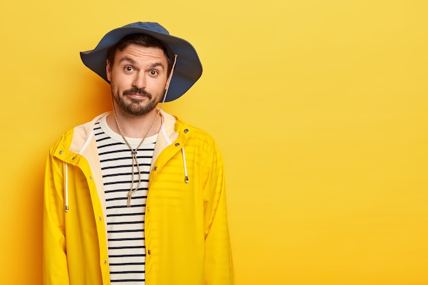 Free photo photo of unshaven man has recreation time, going to fishing during weekend, wears hat and protective raincoat, looks with little surprisement, poses over yellow wall, free space