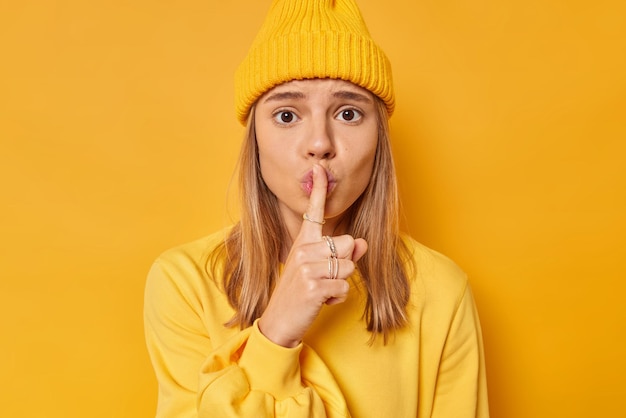 Free Photo photo of worried young female model makes shush gesture presses index finger to lips tells to be quiet asks to keep silence dressed in casual jumper and hat isolated on vivid yellow