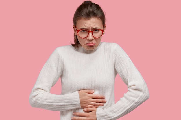 Free photo photo of young attractive dark haired woman purses lips, keeps hands on stomach, feels ill, wears white sweater, isolated over pink studio wall