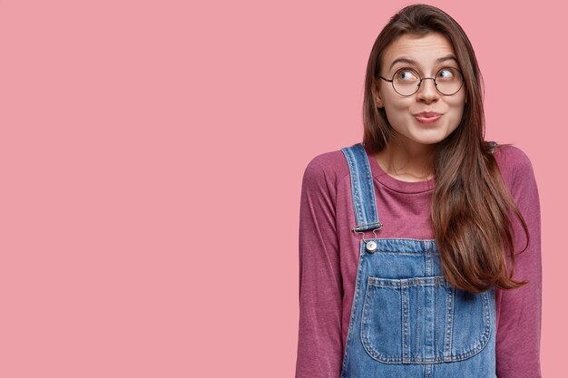 Photo of young dreamy woman with long hair, wears round spectacles, looks happily upwards, has round spectacles, denim overalls