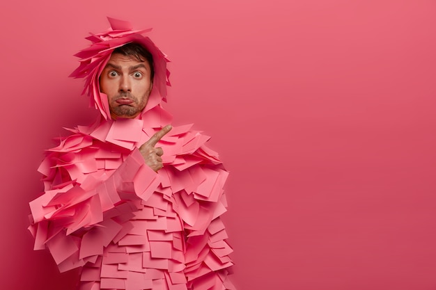 Free photo photo of young man points right displeased, demonstrates bad result to coworkers, purses lips, wears funny paper outfit made of stickers, doesnt like something, shows copy space on pink wall