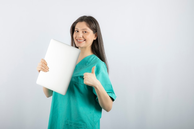 Photo of young nurse with laptop giving thumbs up on white. 