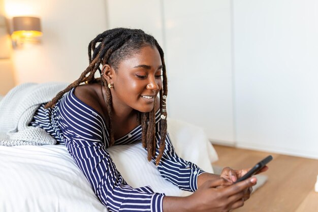 Photo of a young smiling African woman has a video call at night while lying on a bed in her bedroom