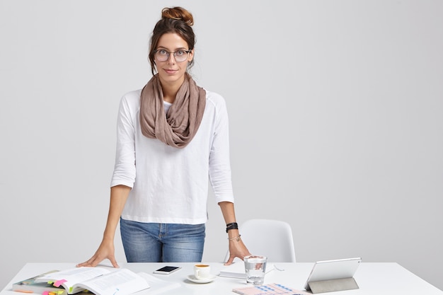 Photo of young teacher in stylish outfit, prepares for seminar or lesson, studies different literary sources, drinks latte, uses tablet for searching information in internet, isolated over white wall