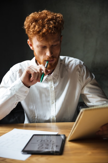 Free Photo photo of young thinking readhead bearded man in white shirt, reading notes, sitting at wooden table