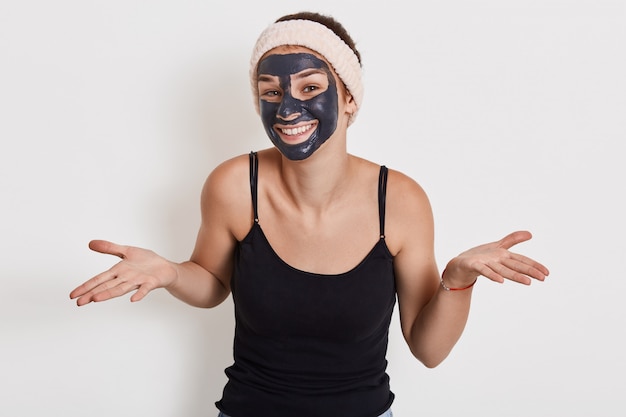 Photo of young woman with charming smile spreading palms aside, shows helpless gesture, wears nourishing facial mask for reducing wrinkles, poses against white wall.