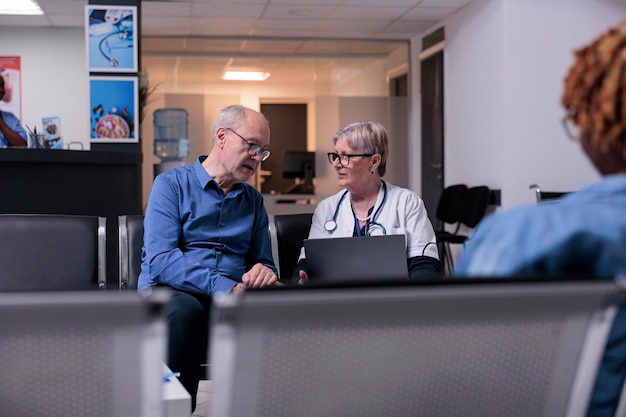 Free photo physician using laptop at checkup visit with old man, sitting in waiting room lobby. general practitioner and patient talking about disease diagnosis and healthcare, consulting adult at health center.