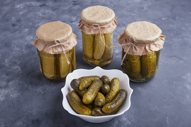 Pickled cucumbers in bowl and glass jars on blue surface.