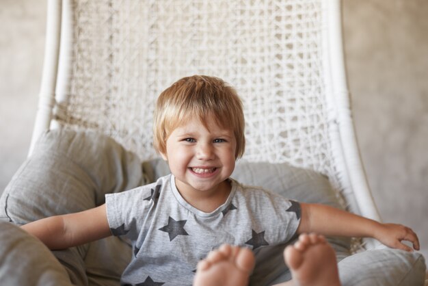 Picture of adorable cheerful little girl with bare feet and short hairstyle, sitting in basket woven chair at home and smiling broadly, feeling excited and impatient while going to watch cartoons