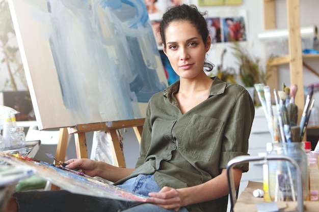 Free Photo picture of attractive professional young caucasian female in casual clothes holding palette and painting knife working on oil painting, mixing colors, having inspired expression on her face