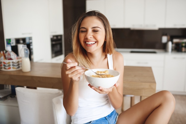 Picture of attractive young girl laughing while eating cornflakes with milk at kitchen