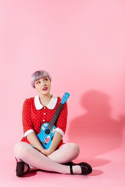 Free photo picture of beautiful dollish girl with short light violet hair wearing red dress holding blue ukulele over pink wall