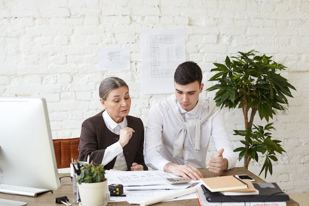 Picture of beautiful experienced elderly woman chief architect helping her skillful young man trainee with architectural project, revising his drawings and pointing at mistakes and drawbacks