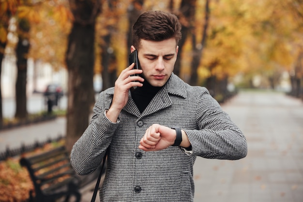 Picture of businesslike man speaking on mobile phone while going on meeting, checking time with watch on hand