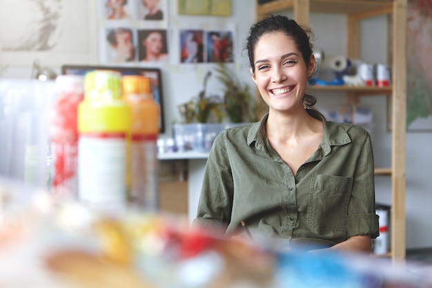 Free Photo picture of charming charismatic young female artist wearing khaki shirt grinning broadly feeling happy about her job and creating process, sitting at workshop, surrounded with painting accessories