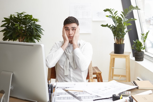 Picture of confused shocked young male designer or architect working on office, feeling stressed and nervous, keeping hands on his head, staring at computer screen, noticing mistake in his drawings