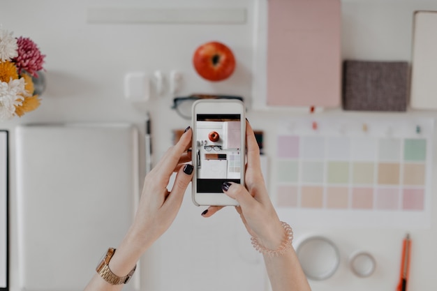 Picture of female hands taking portraits of desktop with stationery, glasses and apple on smartphone