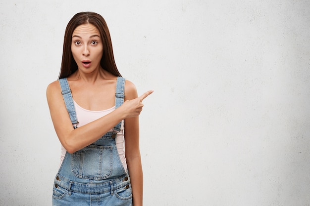 Picture of funny emotional young Caucasian woman with long straight dark hair raising eyebrows and opening mouth in surprise and shock, pointing finger sideways at white blank copy space wall