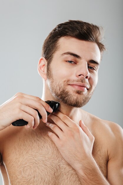 Picture of good-looking adult guy doing hygiene and health procedure with shaving his bristle using trimmer over grey wall