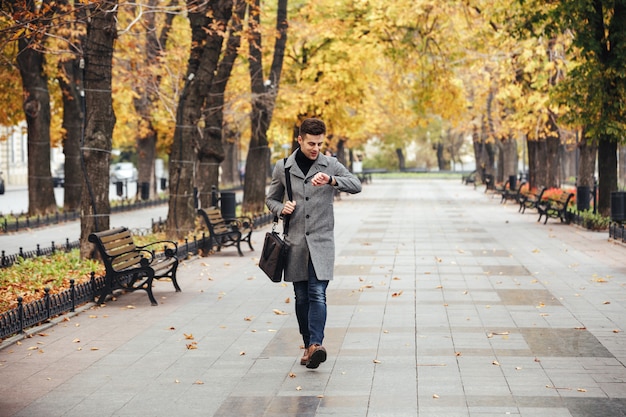 Free photo picture of good-looking caucasian man in coat with bag strolling in city park, and looking at his watch