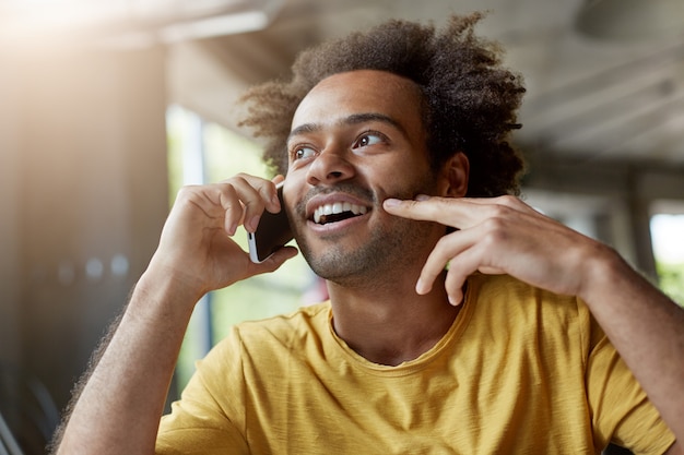 Picture of good-looking happy African man with beard and curly hair smiling cheerfully while talking on mobile phone, having interested look