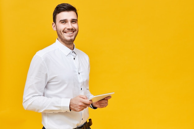 Picture of handsome confident young man in white shirt holding generic digital tablet and smiling broadly, enjoying playing games using online application. Technology, entertainment and gaming