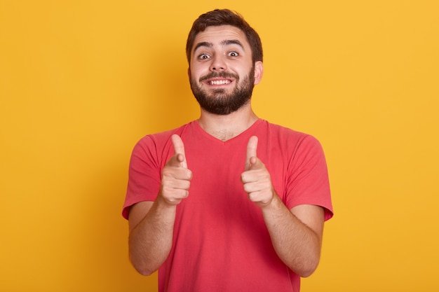 Picture of handsome smiling modern man dresses red casual t shirt showing ok sign with both thumbs, model posing isolated on yellow, bearded young male with happy facial expression.