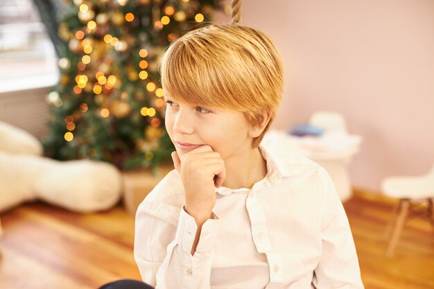 Picture of handsome teenage boy in white shirt having pensive thoughtful look, touching chin, thinking where mother hid New Year's presents, posing in living room with Christmas tree