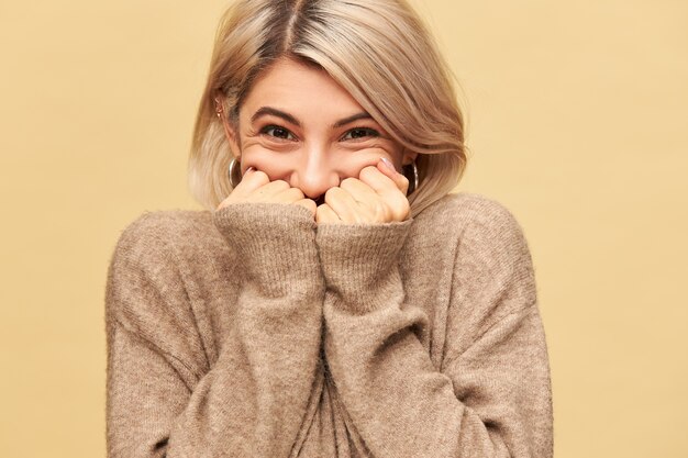 Picture of happy joyful young European woman canât hide her ecstatic emotions, being in good mood, overwhelmed with positive great news, hiding mouth behind hands, wearing long sleeved cozy sweater