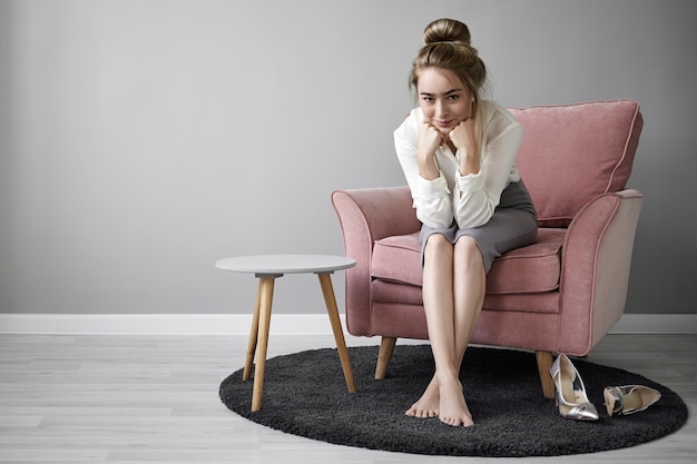 Picture of positive charming young European businesswoman wearing elegant formal clothes sitting in armchair with bare feet on carpet at home, smiling mysteriously, resting chin on her hands