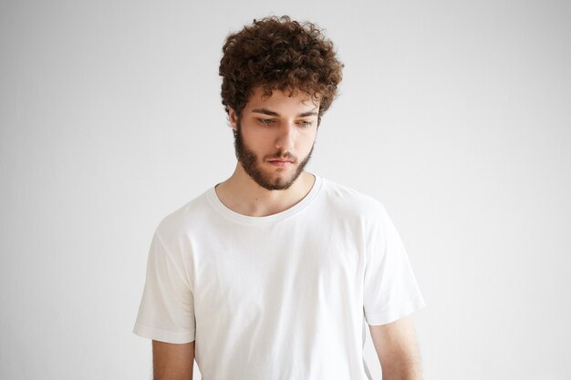 Picture of sad young European male with thick beard looking down having pensive deep in thoughts facial expression, thinking over problems, posing isolated against blank wall