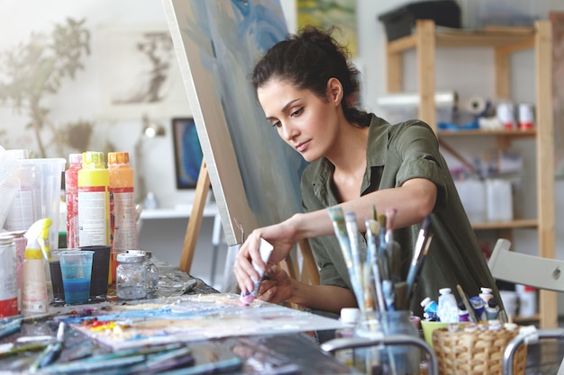 Free photo picture of serious concentrated young caucasian female artist sitting at desk with painting accessories, holding tube of oil paint, mixing colors on palette; unfinished painting on canvas near her