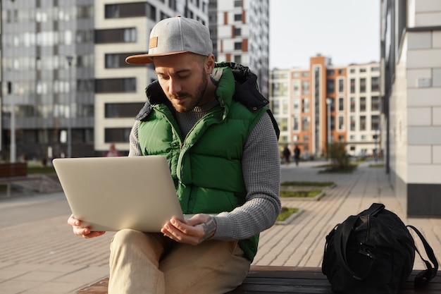 Free photo picture of serious focused young male with stubble surfing internet, using 4g wireless connection on laptop. bearded freelancer working distantly on generic electronic gadget outdoors in cityscape