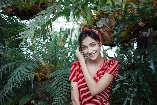 Free photo picture of stylish brunette young european female in fashionable dress and headwear feeling happy and relaxed standing against big green leaves of exotic plants background, smiling broadly at camera