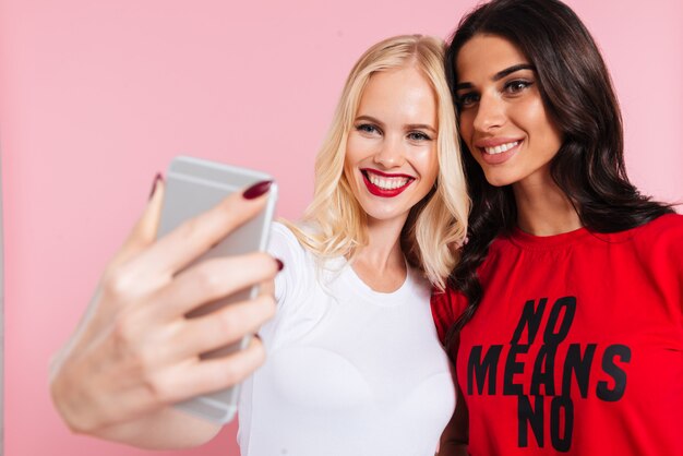 Picture of Two pretty happy women making selfie on smartphone over pink 