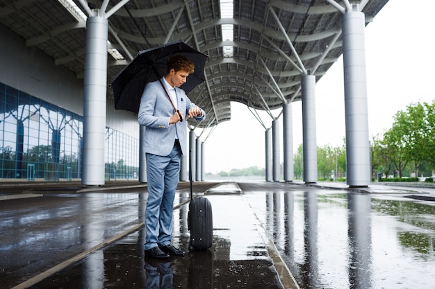 Picture of  young redhaired businessman holding black umbrella in rain and looking on watch