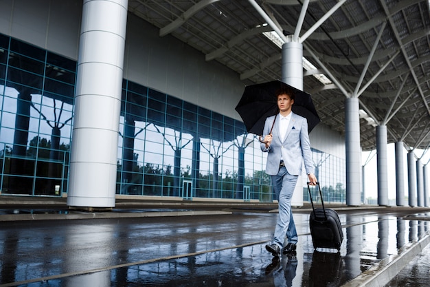 Free photo picture of  young redhaired businessman holding black umbrella and suitcase walking in rain at station