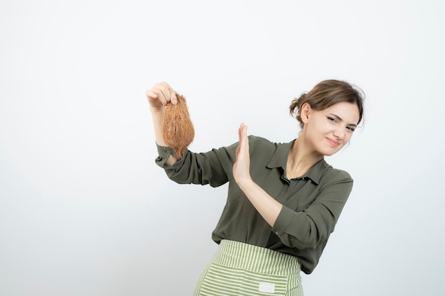 Picture of young woman in apron holding a coconut against white wall . High quality photo