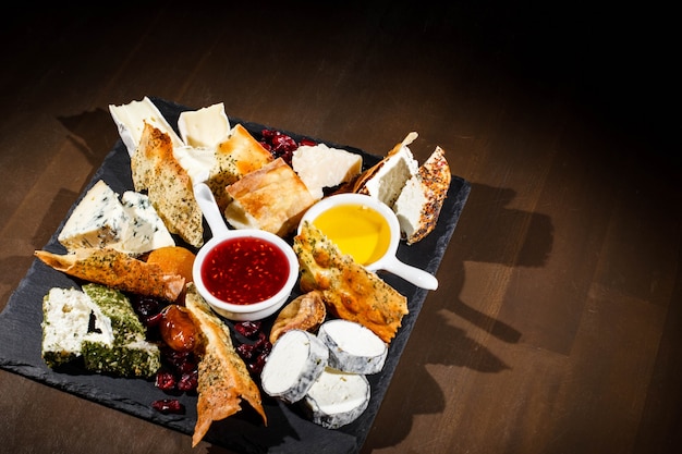 Pieces of camembert,  blue cheese, cheddar lie on black plate with sauces in white bowls