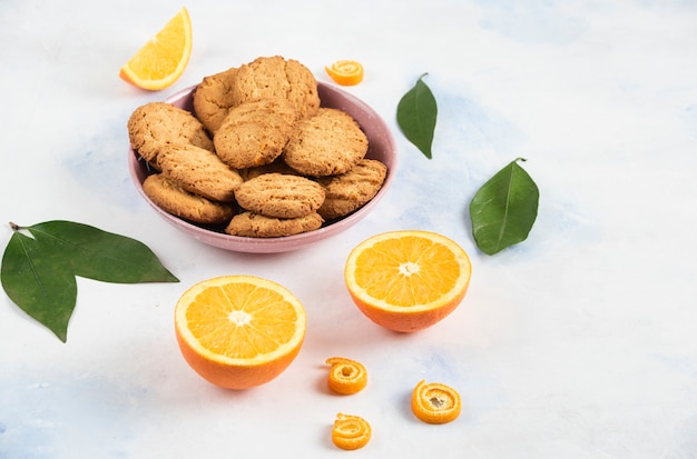 Pile of cookies in pink bowl and half cut oranges with leaves over white table.