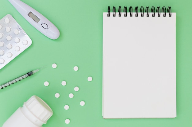 Free Photo pills; thermometer; syringe; plastic bottle near the spiral notepad on green background