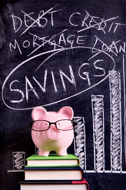 Pink piggy bank with glasses standing on books next to a blackboard with savings message.  