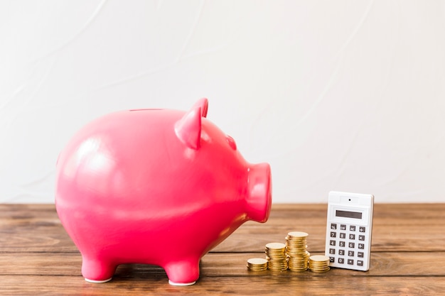 Pink piggybank besides calculator and stacked coins on wooden surface