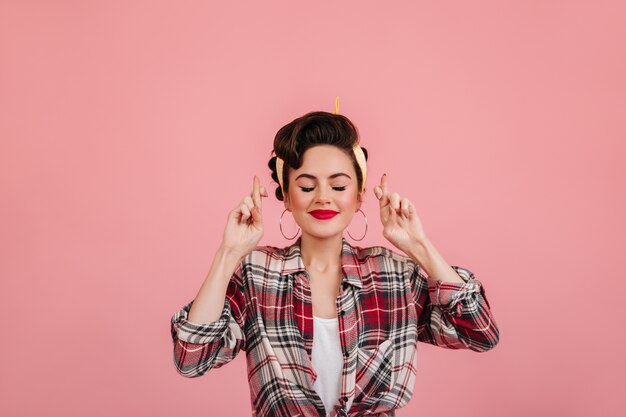 Pinup girl posing with eyes closed and crossed fingers. Cheerful caucasian lady in checkered shirt standing on pink background.