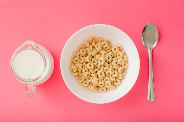 Free photo pitcher of milk and bowl of cereals and spoon on the red background