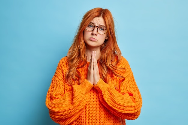 Pitty redhead woman purses lips and keeps hands in pray asks for favor has discontent expression dressed in casual orange jumper.