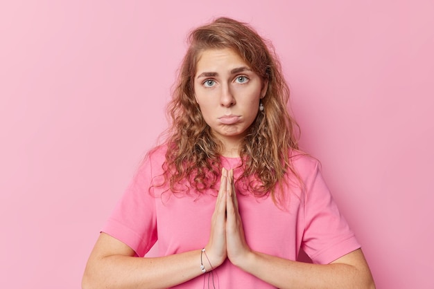 Free photo pity sad blue eyed woman quivers lips with upset expression presses palms together has hopeless begging expression asks for apologize or favor wears casual t shirt isolated over pink background.