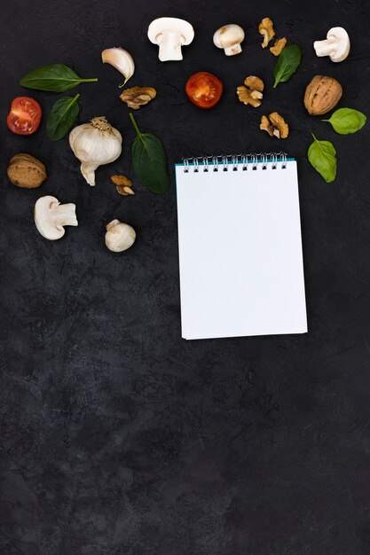 Pizza ingredients over the blank white spiral notepad against black textured background